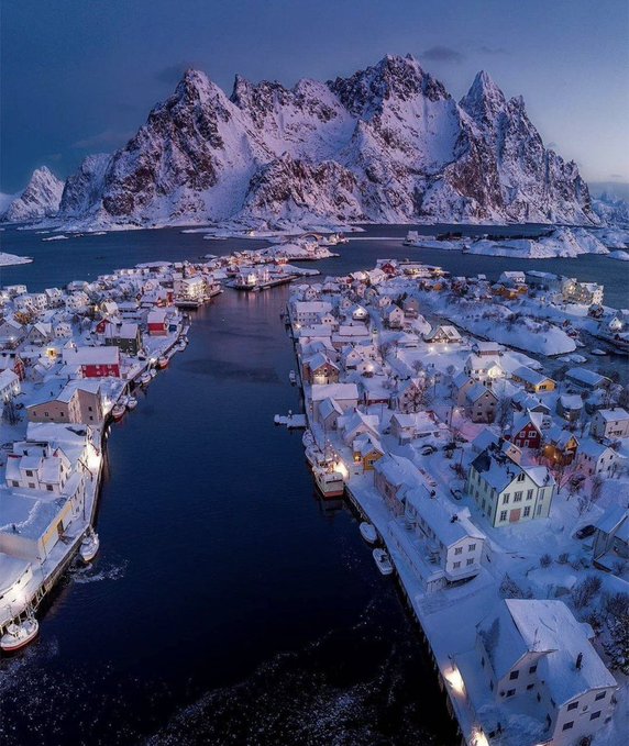Aerial view of a snowy Norwegian fishing village with colorful houses lining a harbor, set against dramatic snow-covered mountain peaks at twilight.