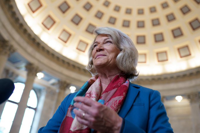 A low-angle portrait of a smiling woman with blonde hair wearing a blue blazer and red scarf, standing inside a domed building.