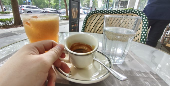 A first-person view of a hand holding a small espresso cup on a saucer, alongside a tall glass of orange juice and a water glass at an outdoor café table.
