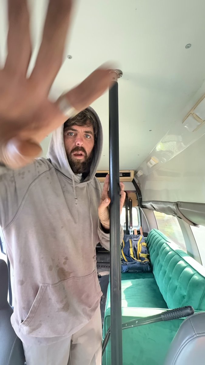 A bearded man wearing a dirty gray hoodie stands inside a vehicle with green seats, holding a metal pole and reaching his other hand toward the camera.