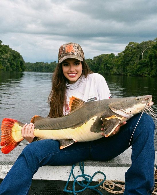 A woman wearing a camouflage cap and white long-sleeve shirt holding a large red-tailed catfish while sitting on a boat on a river surrounded by jungle.