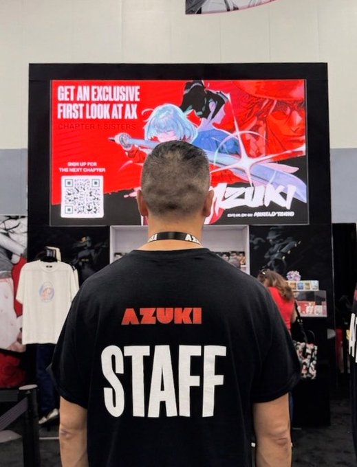 A staff member wearing an "AZUKI STAFF" shirt stands before a digital display promoting "CHAPTER 1 | SISTER 5" for the Azuki webtoon, exemplifying the hard-working Azuki team member being recognized in the caption.