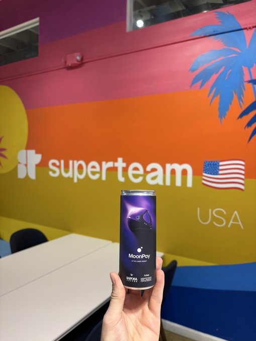 A hand holds a tall purple can of "MoonPay" coffee in front of a colorful "superteam" wall featuring a USA flag and palm tree graphics, perfectly matching the "MoonPay coffee in the wild" caption.