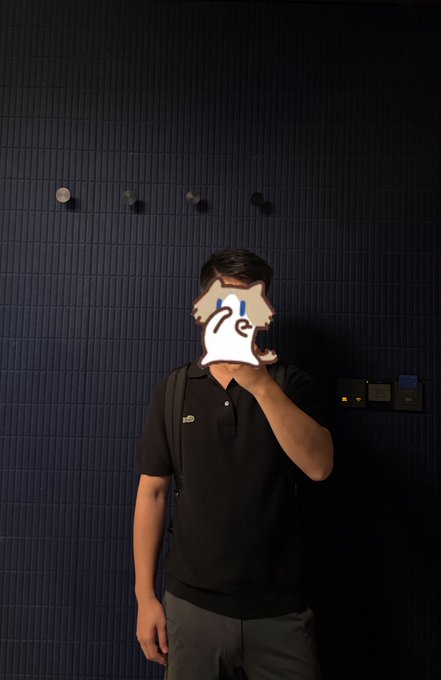 A man in a black polo shirt stands against a dark tiled wall, his face obscured by a ghost emoji, with coat hooks and light switches visible behind him—capturing the relatable sigh of someone reluctantly heading to work while holding onto sky-high crypto dreams.