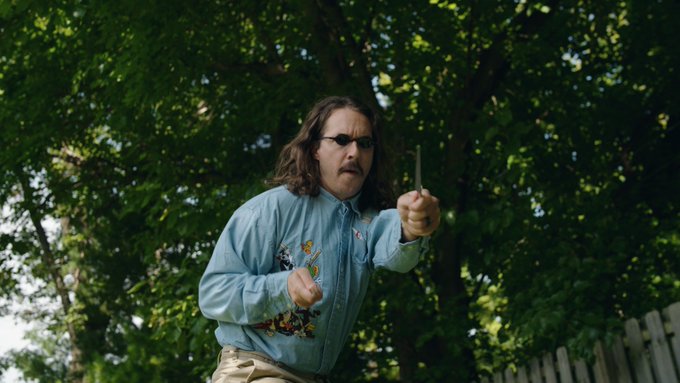 A man with long brown hair, a mustache, and sunglasses, wearing a light blue button-up shirt, stands outdoors holding up a metal object while looking toward the camera with trees in the background.