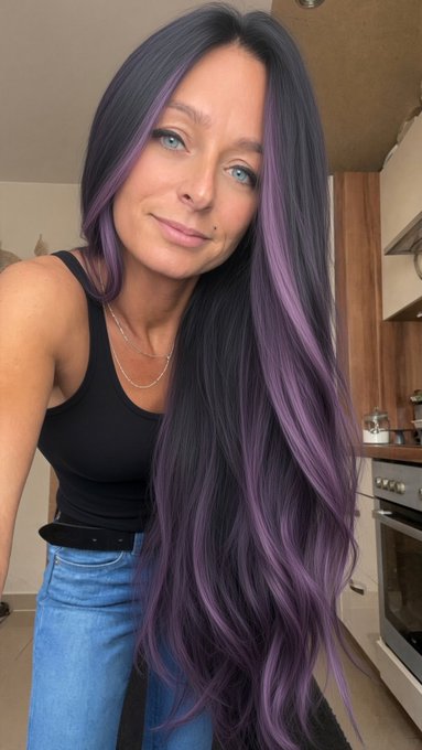 A selfie of a woman with long dark hair featuring purple highlights, wearing a black tank top and blue jeans, standing in what appears to be a kitchen.