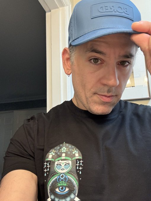 A man wearing a blue 'AESOP' cap and a black t-shirt featuring a graphic of a matryoshka doll with a green alien head and an eye inside.