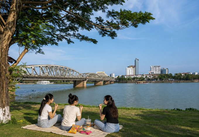 Three women sit on a picnic blanket under a tree on a grassy riverbank, eating snacks with a steel arch bridge, river, and modern city skyline visible in the background.