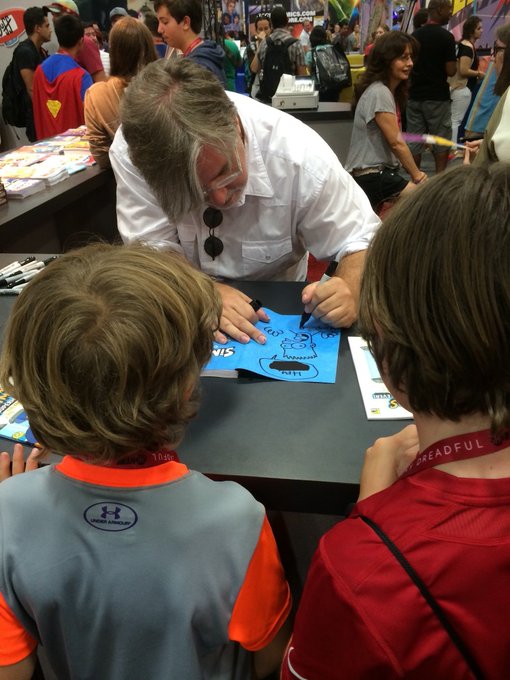 A man in a white shirt signs a blue card reading "Thanks" with a pen for two young fans at what appears to be a comic convention booth. The image captures the personal connection between creators and collectors that the post celebrates, showing how conventions become meaningful spaces for fans to interact with artists whose work holds personal significance.
