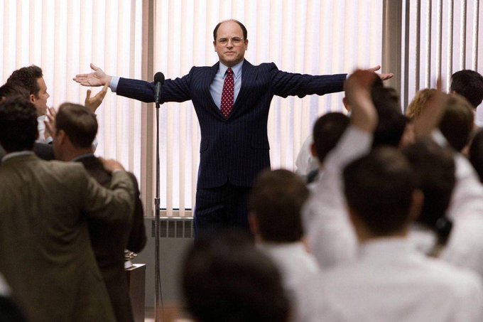 A scene featuring actor Jason Alexander (as George Costanza) in a pinstripe suit standing with arms outstretched while surrounded by an applauding audience.
