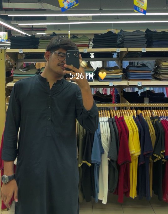 A young man in a black kurta takes a mirror selfie in a clothing store, with a timestamp of "5:26 PM" and a yellow heart emoji overlaid on the image, showing him browsing among shelves of folded garments and racks of colorful polo shirts.