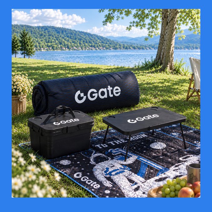 A picnic setup by a lake features a black "Gate" branded blanket, storage box, and folding table.