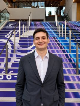 Portrait of a man in a dark suit jacket and white shirt standing in front of a staircase decorated with a blue and purple graphic featuring a scientist character.