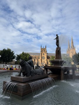 The Archibald Fountain in Sydney sits in the foreground with cascading water features, while St Mary's Cathedral's Gothic spires rise against a cloudy sky in the background.