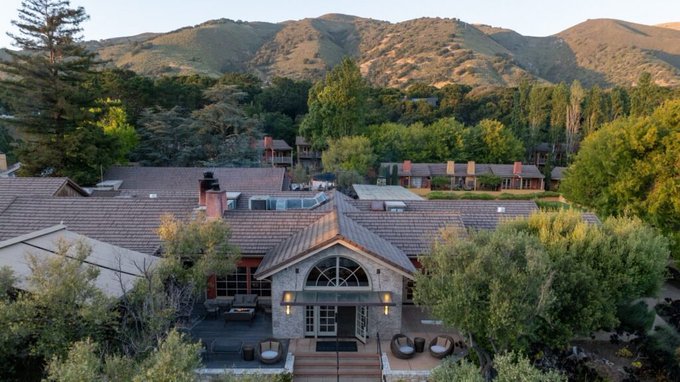 An aerial view of a Tuscan-style villa nestled in the Carmel Valley hills, surrounded by lush greenery and mountains, with a courtyard and outdoor seating area in the foreground—matching the description of the wedding venue for Leopold, where guests gather for a European garden-style celebration.