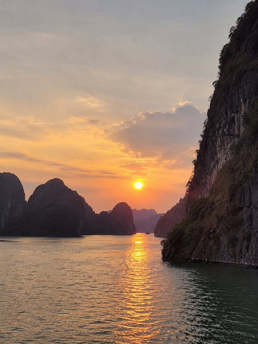 A breathtaking sunset over Halong Bay's limestone karsts, with the golden sun casting a shimmering reflection across calm waters between towering cliffs—capturing the serene, slow-paced weekend energy the post wishes its followers.