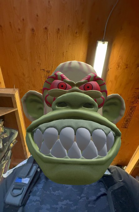 A person wearing a green monkey-like mask with red tribal markings and large teeth, standing in a wooden interior space.