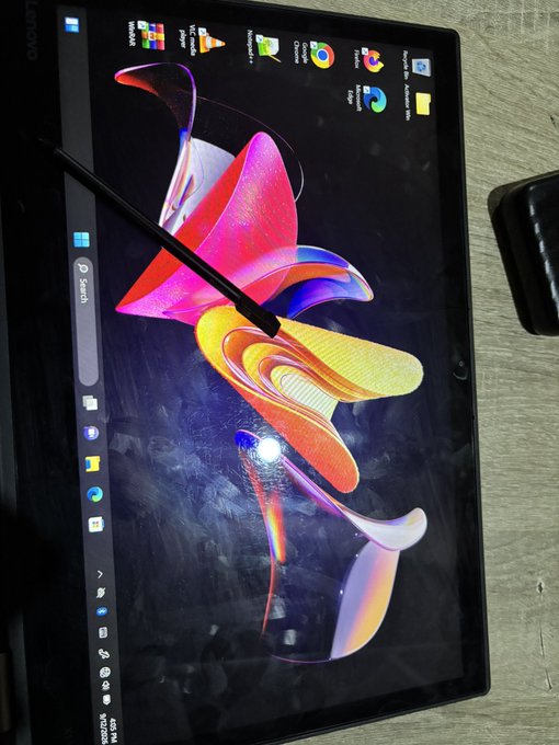 A Lenovo laptop sits open on a desk displaying its Windows desktop with various application icons in the taskbar, alongside a stylus resting on the screen.