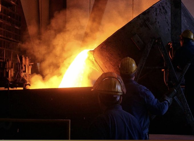Two industrial workers in hard hats observing molten metal pouring from a large furnace in a steel mill.