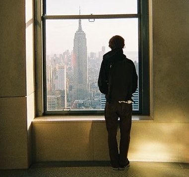 A person stands silhouetted against a window, looking out at the New York City skyline with the Empire State Building prominently visible.