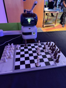 A white humanoid robot with a screen for a face plays chess at a table, viewed across an electronic chessboard with white pieces set up.