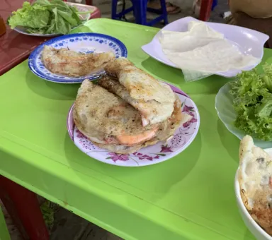 A green plastic table displays plates of food including what appears to be Vietnamese-style pancakes or wraps, accompanied by fresh lettuce and herbs for wrapping.