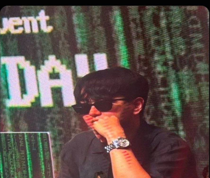 A person wearing sunglasses and a silver watch, sitting in front of a screen displaying green matrix-like code.