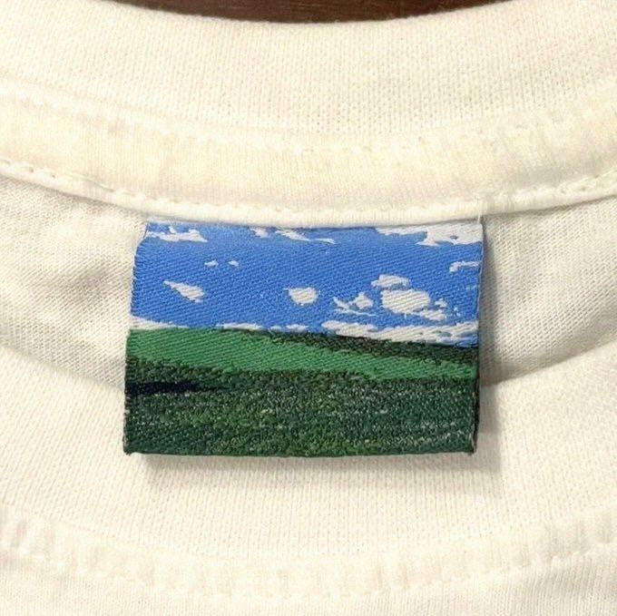 A close-up of a woven clothing label on a white garment depicting a scenic landscape with a blue cloudy sky and green hills/grass.