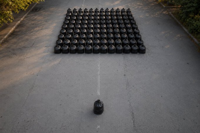 An overhead view of numerous black garbage bags arranged in a grid pattern on an asphalt surface, with a single bag positioned apart at the bottom.