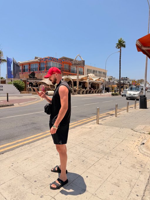A man in a red cap and black tank top stands on a sunny street looking at his phone, seemingly unaware of the excitement building around him. With the caption "A big Crypto pump is coming. GET READY," the image suggests he's checking market signals or crypto updates, ready to act on the anticipated surge.