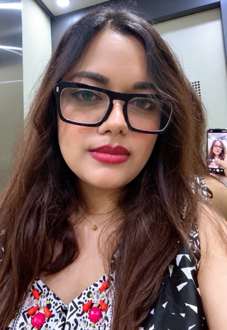 A selfie of a woman wearing black rectangular glasses and a patterned top with red accents, holding a phone.