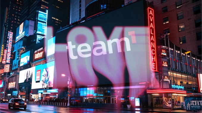 A nighttime cityscape featuring a large illuminated billboard displaying the word 'team1' with Times Square and the 'Avalanche' sign visible nearby.