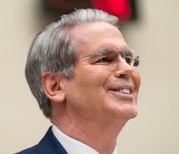 Scott Bessent, the U.S. Treasury Secretary, is shown smiling and looking upward in a close-up portrait, wearing rimless glasses and a dark suit. The image accompanies a critical post highlighting his past hedge fund losses and his current role in influencing cryptocurrency markets through monetary policy.