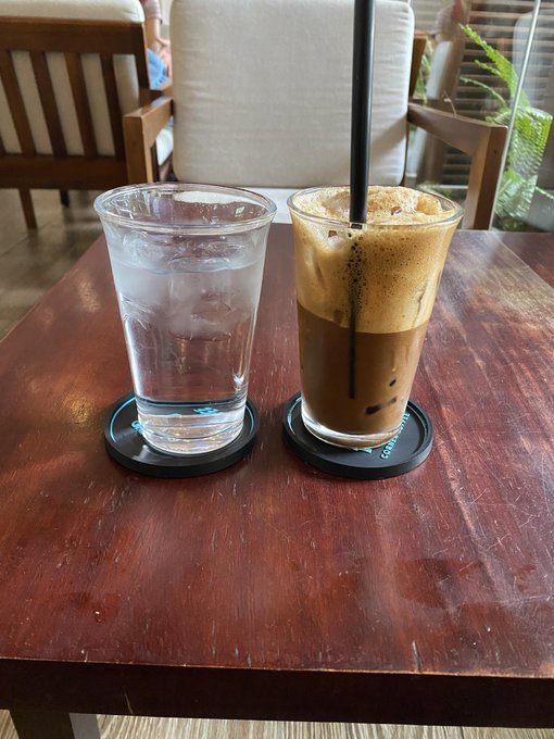 Two drinks on a wooden café table: a clear glass of ice water and an iced coffee in a tall glass with a black straw, sitting on branded coasters.