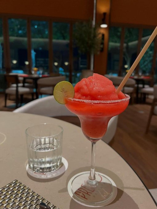 A frozen red margarita-style drink served in a margarita glass with a salted rim and lime garnish, accompanied by a glass of water on a white table.