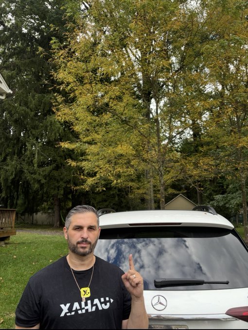 A man wearing a black 'X.AHAU' t-shirt stands in front of a white Mercedes SUV, pointing his finger upward.