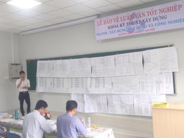 A presentation room with a Vietnamese banner reading "LỄ BẢO VỆ LUẬN VĂN TỐT NGHIỆP" shows a man standing before a wall covered in printed charts and diagrams while two seated individuals observe.