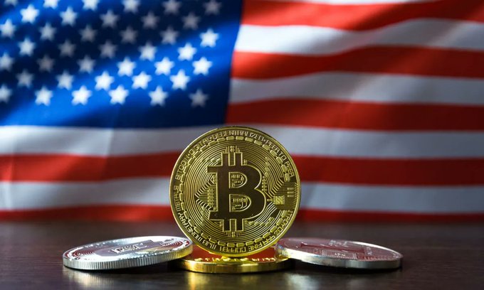 A Bitcoin token stands upright on a wooden surface in front of an American flag, with two other coins lying flat beside it.