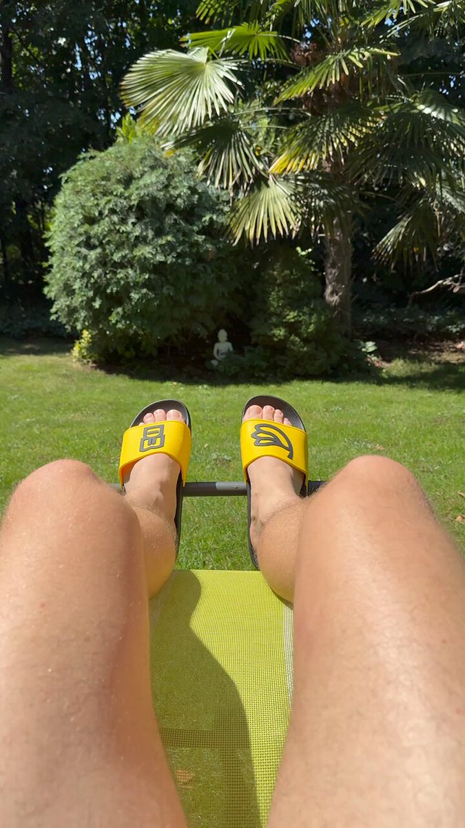 The image shows a person wearing yellow slippers, lounging outdoors in their backyard. The person's legs are crossed over each other while they relax on a green lawn chair. A potted plant can be seen nearby, adding to the outdoor atmosphere of the scene. The photo is captioned with "Unbothered. Moisturized. Happy. Earning SOL yield with @sanctumso on my @solanamobile Seeker." This suggests that the person in the image is enjoying their time outdoors and taking care of themselves by staying hydrated, wearing comfortable slippers, and possibly engaging in outdoor activities like gardening or spending time in nature.