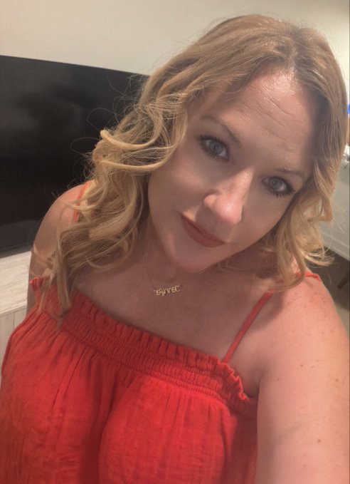 A woman with wavy blonde hair wearing a red off-the-shoulder top takes a selfie, sporting a gold necklace that reads "Byrd." The cheerful, fresh-faced look pairs perfectly with the upbeat "Gm Happy Monday" greeting, capturing a bright start to the week.
