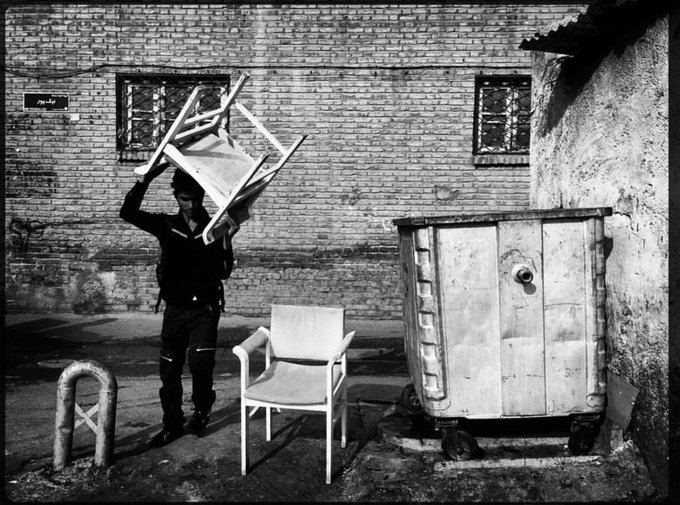 A black-and-white photograph of a young man carrying a wooden chair over his head down a narrow alleyway, with another white plastic chair and a large metal dumpster nearby against a brick wall.
