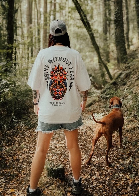 A woman stands on a forest trail with her dog, wearing a t-shirt that reads "SHINE WITHOUT FEAR" and "WORN TO DEATH SINCE MMXXIV" alongside a sun graphic—perfectly embodying the post's message of taking up space and wearing the loud tee while following an unconventional path.