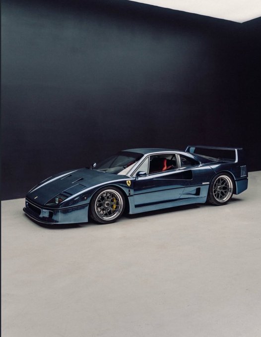 A scale model of a dark metallic blue Ferrari F40 sports car with iconic rear wing sits on a light gray surface against a dark backdrop, capturing the essence of a high-performance exotic supercar that represents extreme wealth and exclusivity, tying into the post's message about how holding $1,000 in the right meme coin could theoretically yield enough returns to afford such a luxury vehicle.