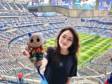 A woman smiles while holding up a plush mascot toy inside MetLife Stadium, with the partially visible "FIFA World Cup 2026" signage on the jumbotron in the background. Her excitement about wanting the limited-edition World Cup Fire Treasure merchandise aligns perfectly with the stadium setting as she hopes to score the exclusive collectible.