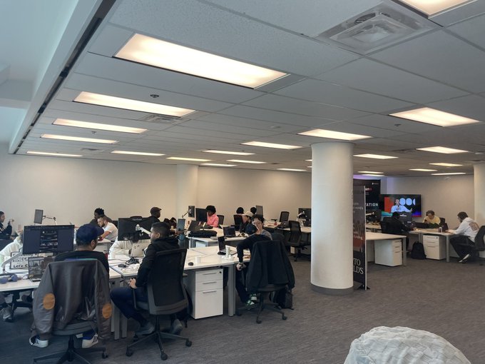 An office workspace with several employees working at desks with computers in a brightly lit room with white pillars.