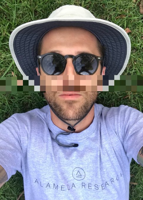 A person wearing a gray 'ALAMEDA RESEARCH' t-shirt, a bucket hat, and sunglasses lies on grass, with their eyes blurred out.