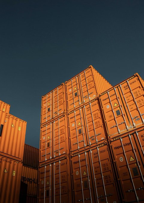 A low-angle photograph of stacked orange shipping containers branded with 'Hapag-Lloyd' against a clear dark blue sky, with one container marked '20 PC-LN.'