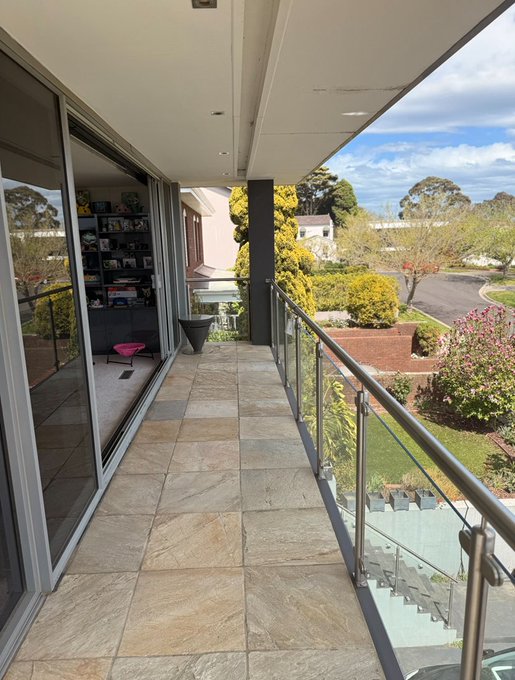 A sunlit balcony with stone tile flooring, glass railings, and sliding doors opens to a view of a tree-lined suburban neighborhood. The image pairs with the "Can I get a GM?" post, offering a serene morning vista perfect for a cheerful greeting.