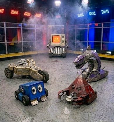 The image features four toy robots, each with different colors and designs, sitting on a gray floor. The first robot has blue eyes and a yellow body, while the second one has red eyes and a purple body. The third robot has orange eyes and a white body, and the fourth one has black eyes and a brown body. These robots are positioned in front of a glass wall that separates them from an audience watching their performance on stage.