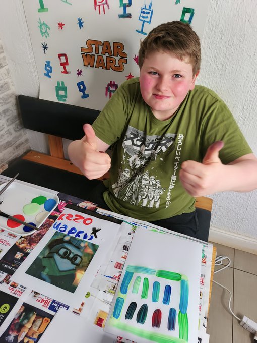 A young boy in a green Star Wars shirt giving two thumbs up, seated at a table with artwork and a tablet displaying a drawing app with green and blue brush strokes.